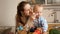 Cute baby boy sitting on kitchen table and playing with cookware while mother feeding him with porridge. Concept of little chef,