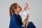 Cute baby in his mother`s arms on a gray background in the studio. A mother in denim clothes lovingly embraces a child. The