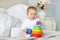 Cute baby playing with colorful rainbow toy pyramid sitting on bed in white sunny bedroom. Toys for little kids.