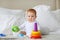 Cute baby playing with colorful rainbow toy pyramid sitting on bed in white sunny bedroom. Toys for little kids.