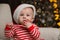 Cute baby in Santa hat and bright Christmas pajamas eating candy cane at home