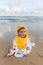 Cute baby wearing a yellow cartoon bathrobe sitting and playing on the beach near the sea