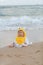 Cute baby wearing a yellow cartoon bathrobe sitting and playing on the beach near the sea