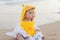 Cute baby wearing a yellow cartoon bathrobe sitting and playing on the beach near the sea