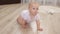 Cute barefoot baby infant girl is crawling on wooden floor in living room at home, looking at camera, smiling and change