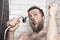 Cute bearded man singing in the bathroom using the shower head with flowing water instead of a microphone