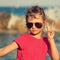 Cute beautiful kid girl in fashion sunglasses showing v sign making the selfie on blue sea and sky background. Toned closeup port