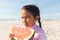 Cute biracial girl holding large fresh watermelon slice looking away at beach on sunny day