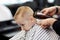 Cute blond baby boy with blue eyes in a barber shop having haircut by hairdresser. Hands of stylist with tools. Children fashion i