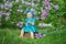 Cute blond baby girl enjoying time on a awesome place between lilac syringe bush.Young lady with basket full of flowers dressed in