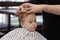 Cute blond serious baby boy with blue eyes in a barber shop having haircut by hairdresser. Children`s fashion.