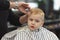 Cute blond smiling baby boy with blue eyes in a barber shop having haircut by hairdresser. Hands of stylist with tools. Children f