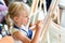Cute blond smiling girl painting on easel in workshop lesson at art studio. Kid holding brush in hand and having fun