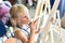 Cute blond smiling girl painting on easel in workshop lesson at art studio. Kid holding brush in hand and having fun drawing with