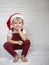 Cute blond toddler boy in white T-shirt, stylish red plaid overalls and Santa hat, barefoot, sits on light background