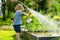 Cute blond toddler with a watering pot outdoors in the garden. Kid helping parents with gardening in the backyard in bright sunny