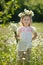 Cute blond young girl child stylish dressed in white shirt wearing wreath of daisy chamomiles posing on meadow of forest wild mayw