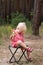 Cute blonde girl sits on picnic chair in the forest. Vertical frame