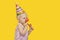 Cute blonde girl is wearing party hat and red clown nose and holding whistle. Yellow background. Copy space