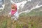 Cute blonde girl wearing pink scarf surrounded with spectacular alpine scenery exploring mountain meadow.
