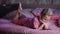 Cute blonde small girl dressed in pink pyjamas is lying in bed and watching cartoons with smartphone. Toddler female kid