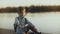 Cute boy child sits with a stick near sunset lake. Portrait of Caucasian child playing angry wild pirate on a pier. 4K.