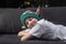 Cute boy in christmas elf hat is resting on the sofa with closed eyes on the background of Christmas tree. Anticipation of holiday