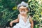 Cute boy in hat and casual clothes sitting around tomatoes ang looking at camera, kid model posing in garden