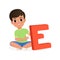 Cute boy sitting with crossed legs and holding big letter E. Cartoon character of little kid. Fun educational game. Flat