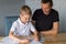 A cute boy is sitting with his dad at the table and collecting origami. Holds a white sheet of paper