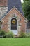 Cute brick and rock cottage with gabled entrance and arched door with bright green lawn and fall wreath on door.