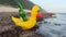 A cute bright yellow inflatable duck with a beer bottle inside it on ocean reef on white sand wild beach in tropical