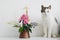 Cute cat sitting at summer flowers bouquet in vintage vase on white  table. Home decor and pet. Curious kitty playing at stylish