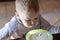 Cute caucasian baby boy with blond hairs blows into the porridge at the table, in sunny morning.