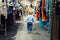 Cute caucasian blond toddler boy walking alone at clothes retail store between rack with hangers. Baby discovers adult shopping