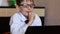 A cute caucasian boy in glasses 7 years old is studying at home doing homework sitting at a table in front of a gadget. Children a