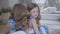 Cute Caucasian brunette girl in blue dotted dress smiling as listening to twin sister whispering on her ear. Joyful