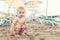 Cute caucasian toodler boy squatting alone on sandy beach between chaise-lounge. Adorable happy child having fun playing at