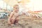 Cute caucasian toodler boy squatting alone on sandy beach between chaise-lounge. Adorable happy child having fun playing at