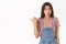 Cute cheerful european brunette girl in overalls, t-shirt, pointing thumb left, folding lips curious and entertained