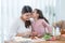 Cute child daughter kissing Asian mother on cheek while mom preparing breakfast in kitchen, peeling apples, milk, bread,