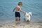 Cute child play with little dog in at seashore. Boy stand in sea water near white dog. Friends going to swim together