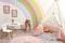 Cute child`s room interior with stylish furniture, toy tent and rainbow art on wall