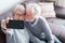 Cute couple of two seniors married taking a selfie together sitting on the sofa at home - mature man kissing his wife while she`s