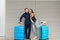 Cute couple with two suitcases is standing on gray striped background. She has long hair, glasses, yellow sweate