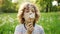 A cute, curly-haired boy blowing a dandelion. Summer glade, sun rays. Children Day. Dynamic video