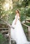 A cute curly woman in a white wedding dress with a wedding bouquet and wreath in her hair standing back to the camera in nature.