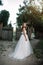 A cute curly woman in a white wedding dress with a wedding bouquet and wreath in her hair standing back to the camera in nature.