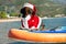 Cute dachshund dog in sunglasses for pets with polarizing lenses and Santa costume is standing on SUP board. Outdoor