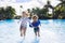 A Cute diverse boy and little girl running and splashing together holding hands in the large swimming pool resort while on a famil